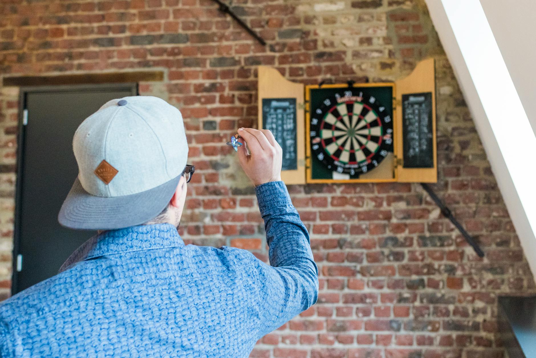 back view photo of man in blue dress shirt and gray hat playing darts